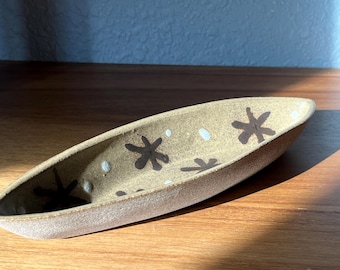 May include: A ceramic serving dish in the shape of a boat with a speckled brown glaze. The dish is decorated with white dots and black star-shaped designs.