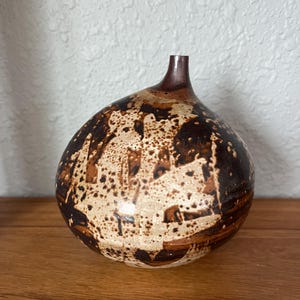 May include: A handcrafted ceramic vase with a rounded body and a narrow neck. The vase features a speckled design in shades of brown and cream, creating a unique, organic aesthetic. The vase is displayed on a wooden surface.