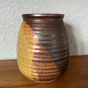 May include: A rounded ceramic vase featuring a ribbed, horizontal texture. The exterior displays a two-tone glaze with a warm, speckled mustard yellow on one side and a deep, dark brown on the other, meeting in a diagonal transition across the body.