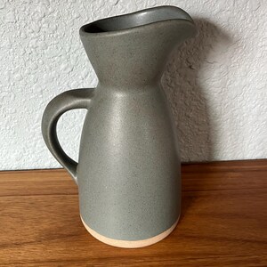 May include: A ceramic pitcher with a speckled dark gray glaze on the upper body and a smooth, unglazed light tan base. The vessel features a curved handle on the side and a wide, flared pouring spout at the top, creating a functional silhouette.