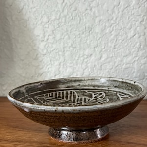 May include: A small, round, footed ceramic bowl with a dark brown, speckled exterior glaze. The interior features a contrasting cream-colored glaze with a hand-drawn, white line-art leaf pattern etched into the center of the basin.