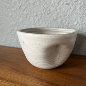 May include: A small, off-white ceramic bowl with a speckled finish and a distinct thumb indentation on the side. The bowl features horizontal ridges along its exterior surface, a wide circular opening, and a smooth, rounded base design.