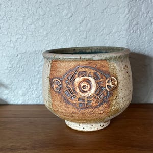 May include: A handcrafted ceramic bowl with a beige and brown color scheme. The bowl features a decorative geometric design on the front, with a circular pattern in the center. The bowl has a rounded shape and a small, raised base. The rim is a darker shade of brown.