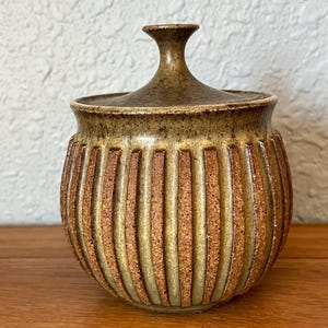 May include: A ceramic jar with a lid, featuring vertical ribbed detailing. The jar has a rounded shape with a brown and tan color scheme. The lid has a small handle. The jar is sitting on a wooden surface.