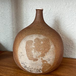 May include: A brown ceramic vase with a rounded body and a narrow neck. The vase has a textured surface and a lighter-colored circular design on the front. The rim of the neck is white. The vase is sitting on a wooden surface.