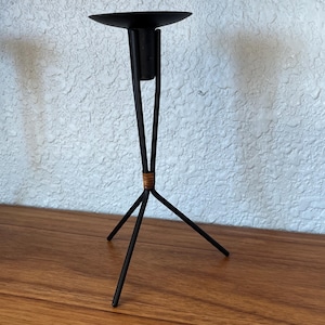 May include: A black metal candle holder with a tripod base and a circular top. The candle holder has a minimalist design with a woven detail near the base of the stem. The candle holder is approximately 8 inches tall.