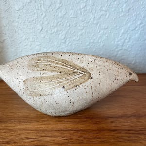 May include: A speckled, ceramic bird sculpture in a neutral color palette. The bird has a stylized wing design in shades of brown and a small beak. The sculpture is placed on a wooden surface.