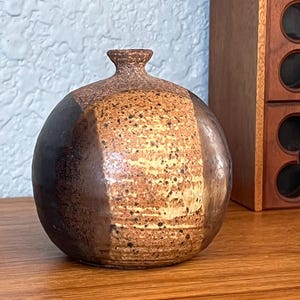 May include: A handcrafted ceramic vase with a rounded body and a narrow neck. The vase features a speckled, textured surface with bands of brown and tan hues. The design is simple and elegant, suitable for displaying flowers or as a decorative piece.