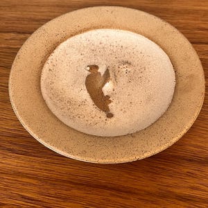 May include: A handmade, beige ceramic coaster with a raised center. The center features a brown bird silhouette. The coaster sits on a wooden surface. The coaster is round and has a slightly textured surface.