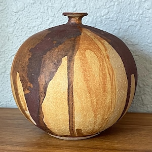 May include: A spherical ceramic vase with a narrow neck and a flat rim. The surface features a textured, matte finish with a color palette of warm tan and deep brown, displaying vertical drip patterns that wrap around the rounded body.