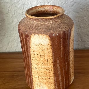 May include: A cylindrical ceramic vase with a speckled cream and tan base, featuring vertical brown glaze drips running down the sides. The rim is rounded and textured with a dark brown finish, and the overall surface has a mottled appearance.