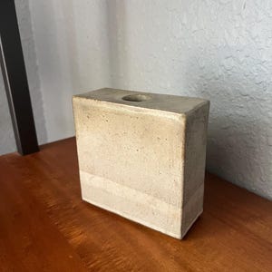 May include: A rectangular, beige ceramic vase with a single circular opening at the top. The vase has a textured surface and is placed on a wooden surface. The vase is a neutral color and has a minimalist design.