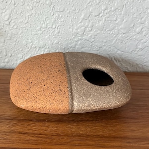 May include: A handcrafted, ceramic decorative object with a bi-color design. The left side is a speckled, reddish-brown, while the right side is a speckled, light gray-brown. It has a rounded shape with a hole on the right side.