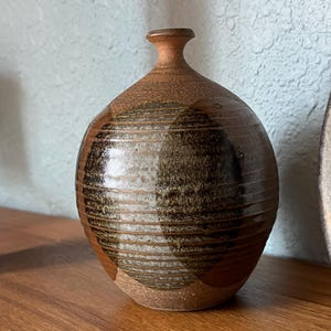 May include: A handcrafted ceramic vase with a rounded body and a narrow neck. The vase features a brown and black striped design around the middle, with a lighter brown base and top. The vase sits on a wooden surface.