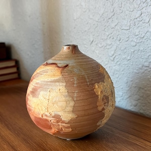 May include: A handcrafted ceramic vase with a rounded body and a narrow neck. The vase features a textured surface with earthy tones of brown, beige, and reddish-orange. The vase is displayed on a wooden surface.