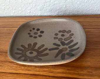 May include: A small, square, ceramic dish with rounded corners. The dish is a muted brown color with darker brown floral designs. The designs include dots and stylized flower shapes. The dish is likely handmade.