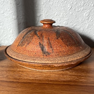 May include: A round, lidded ceramic dish with a speckled, mottled finish in shades of burnt orange, rust, and dark brown. The lid features a small, centered knob handle and a textured, earthy surface with subtle dark streaks radiating from the center.