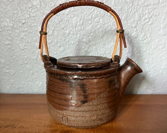 May include: A brown ceramic teapot with a brown and tan speckled glaze. The teapot has a bamboo handle with brown leather accents.