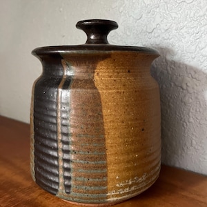 May include: A handcrafted ceramic jar with a lid. The jar features a color scheme of brown, black, and tan. The lid has a small handle. The jar is sitting on a wooden surface.