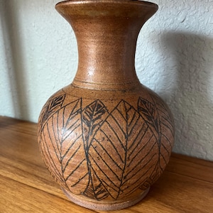 May include: A brown ceramic vase with a flared rim and a rounded body. The surface features a dark brown etched pattern of repeating leaf shapes that wrap around the exterior. The base is unglazed and shows the natural texture of the clay material.