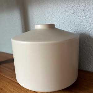 May include: A cream-colored ceramic vase with a cylindrical body and a slightly flared top. The vase has a smooth, matte finish and sits on a wooden surface. The design is simple and modern, suitable for various home decor styles.