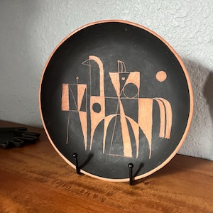 May include: A black ceramic plate with a geometric design in shades of brown. The plate is round with a raised edge and is displayed on a black metal stand. The design features abstract shapes and lines.