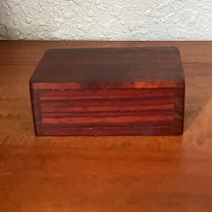 May include: A rectangular wooden box with a smooth, polished finish. The box features a deep reddish-brown color with visible horizontal wood grain patterns running across the front panel, showcasing a natural, dark-toned timber construction.
