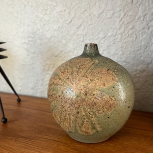 May include: A round ceramic vase with a narrow neck. The vase has a speckled, earthy design in shades of green and brown. The vase is sitting on a wooden surface.