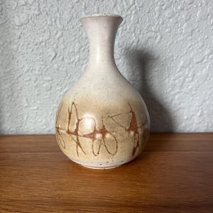 May include: A small ceramic bud vase with a rounded body and a narrow neck. The piece features a speckled off-white glaze on the upper portion, transitioning to a tan base decorated with dark brown, hand-painted abstract line patterns.