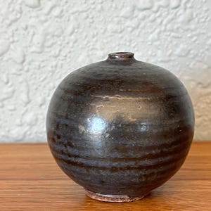 May include: A small, round, dark brown ceramic vase with a narrow neck. The vase has a smooth, glossy finish with horizontal striations and sits on a wooden surface. The vase is approximately 4 inches tall.
