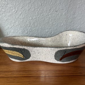 May include: An elongated, kidney-shaped ceramic bowl with a speckled off-white glaze. The exterior features two dark gray oval patches, one containing a yellow leaf motif and the other containing a dark red leaf motif, with a smooth rim.