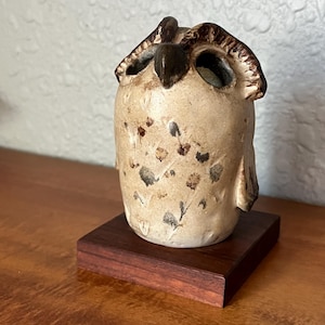 May include: A ceramic owl figurine with a cream-colored body and dark brown accents. The owl has large eye sockets and is perched on a small, dark wooden base. The owl's body is speckled with dark brown spots.