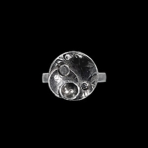 May include: A silver ring with a round, textured design featuring multiple overlapping circles.