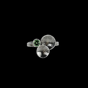 May include: A silver ring with a green gemstone and two textured silver circles. The ring has a unique design with the gemstone set on one side and the circles on the other, all against a black background.