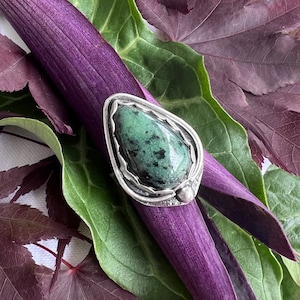 May include: A silver ring with a green gemstone set in a teardrop shape. The ring is on a purple stem with green leaves.