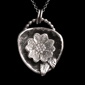 May include: A silver pendant necklace featuring a flower with leaves, set against a textured background. The pendant is attached to a silver chain.
