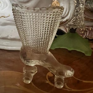 May include: Clear glass shoe-shaped vase with a textured basket weave pattern. The vase has a high heel and is supported by three small feet. The background includes other decorative items.