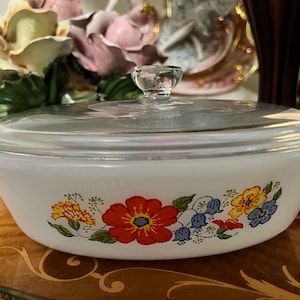 May include: A white, oval casserole dish with a clear glass lid. The dish is decorated with a floral pattern featuring red, yellow, and blue flowers. The lid has a clear, round handle. A vintage kitchenware item.