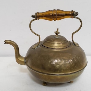 May include: Antique brass kettle with a rounded body, spout, and lid. The kettle features a wooden handle and small feet. The brass has a warm, aged patina, suggesting its vintage. A classic kitchenware item.