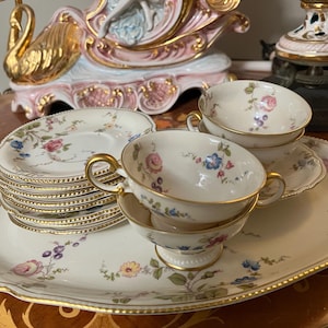 May include: A vintage tea set featuring floral designs in pink, blue, and yellow. The set includes cups, saucers, and a serving tray with gold trim. The cups have gold handles and sit on matching saucers. The tray is oval-shaped.