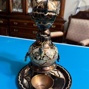 May include: An ornate, antique copper-colored tea kettle with intricate etched designs. The kettle sits on a matching tray with a small cup. The design includes floral and geometric patterns, with a dark brown and gold color scheme.