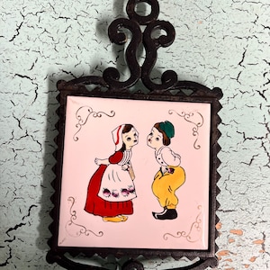 May include: A decorative trivet with a square ceramic tile featuring a cartoon couple in traditional attire. The tile is set in a black wrought iron frame with an ornate design. The background is a distressed, light blue surface.