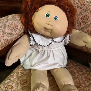 May include: A vintage Cabbage Patch Kid doll with red hair and green eyes. The doll is wearing a white dress with a black trim around the collar and sleeves. The doll is sitting on a floral patterned chair.