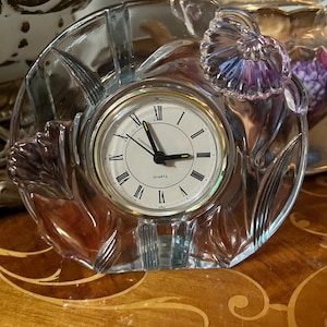 May include: A clear glass quartz clock with a floral design. The clock face is white with black Roman numerals and black hands. The clock is surrounded by a clear glass floral frame.
