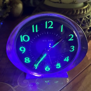 May include: A round Big Ben alarm clock with a metallic silver frame and a black face. The clock features glowing green numerals and hands illuminated by a vibrant purple light. The text Big Ben is printed in white on the center of the clock face.