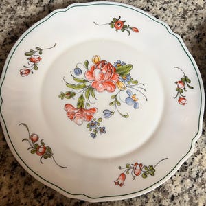 May include: White ceramic plate with a scalloped edge and a green border. The plate features a central floral design with red, orange, yellow, and blue flowers. Additional floral accents are scattered around the rim.
