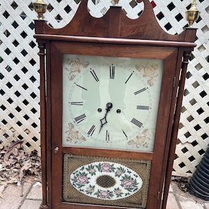 May include: A wooden antique clock with a light-colored face and black Roman numerals. The clock has a decorative floral design on the face and lower panel, with gold-colored accents and finials. The clock is approximately 30 inches tall.