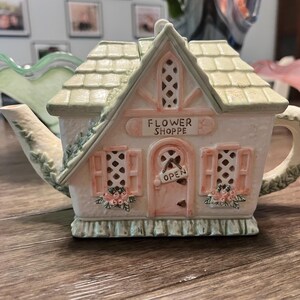 May include: A decorative teapot shaped like a house, with a light pink and green color scheme. The teapot features a roof, shutters, and the text "FLOWER SHOPPE" and "OPEN". The handle and spout have a floral design.
