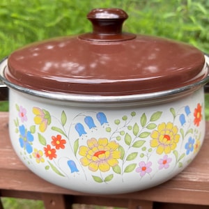 May include: A vintage white enamel pot with a brown lid and handles. The pot is decorated with colorful floral patterns, including yellow, blue, and red flowers, and green leaves. A classic kitchenware item.