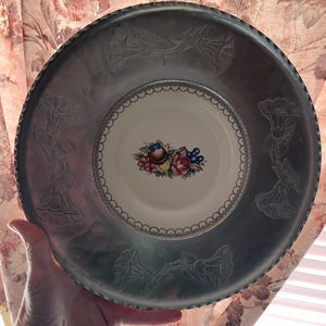 May include: A large, round, silver-toned metal serving tray with embossed floral designs. The center features a white ceramic plate with a floral and fruit design. The tray is held by a hand, showcasing its size and detail.